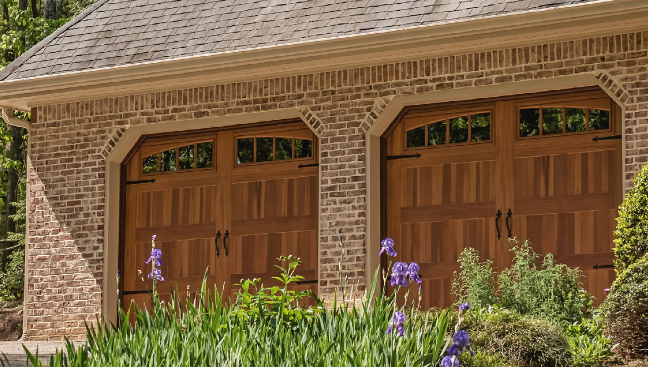 A brick garage with two wooden doors and small windows at the top, surrounded by green plants and purple flowers.