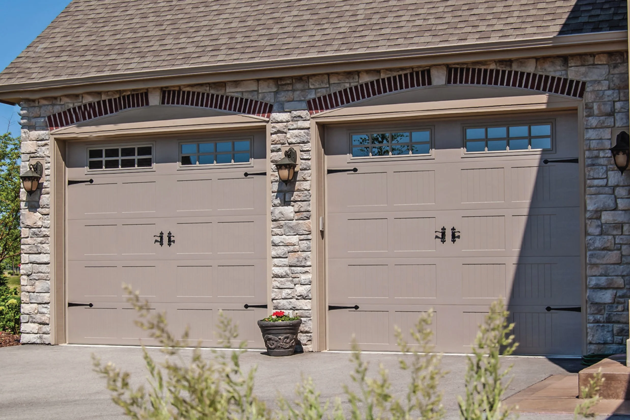 Double garage doors with small windows at the top, surrounded by stone façade and brick accents, with outdoor wall lanterns on each side.