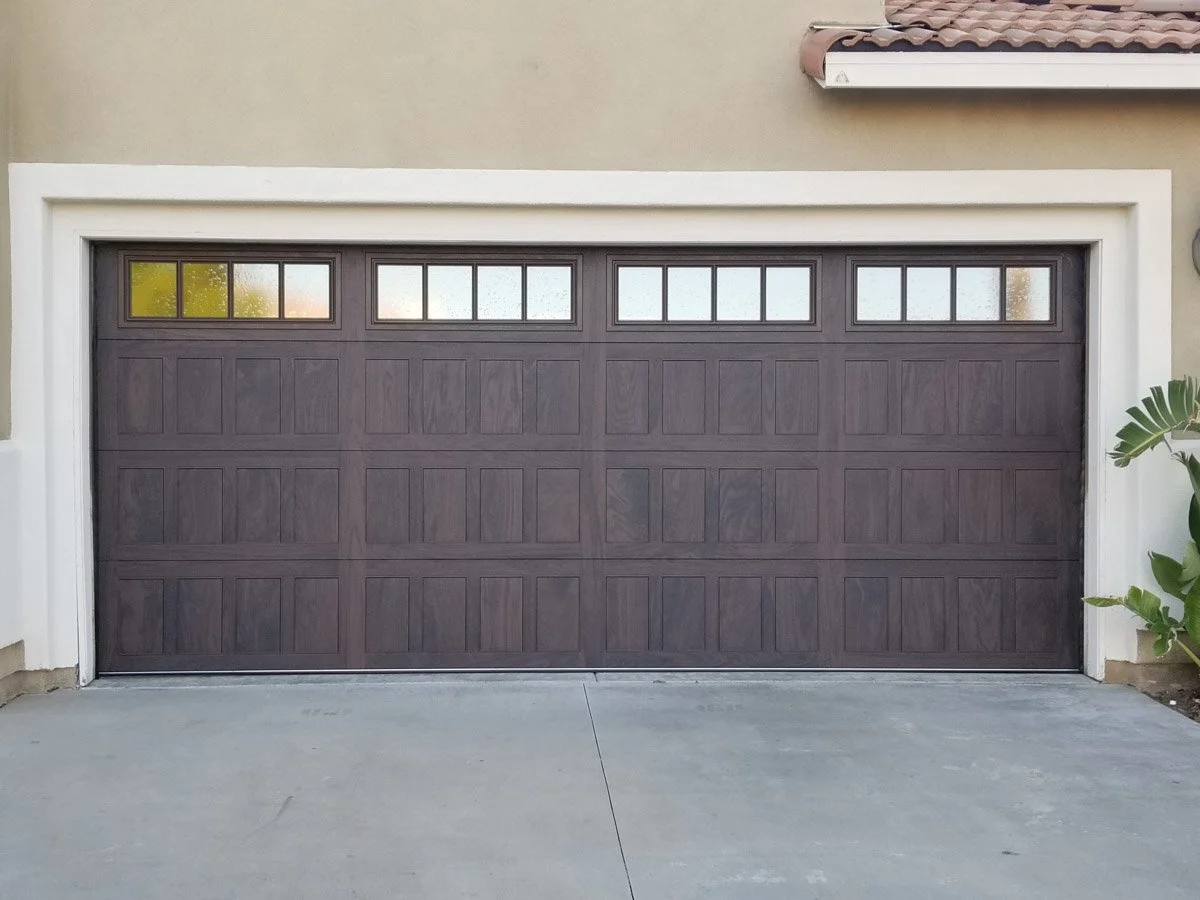 A closed garage door made of wood with rectangular panels, located in a residential house, with a concrete driveway in front and a small green plant on the right side.