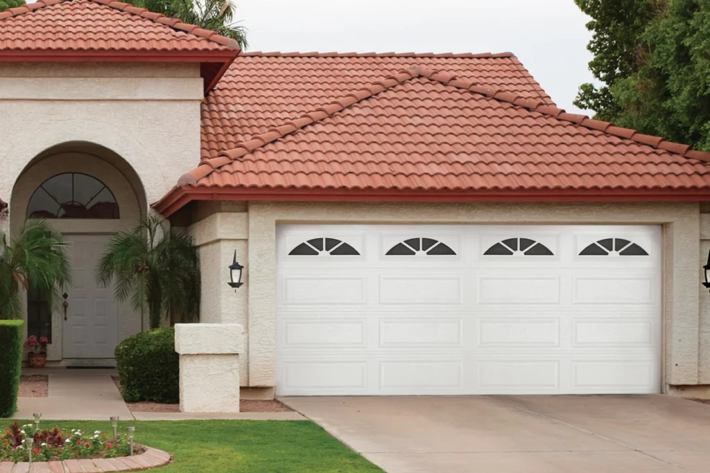 Front view of a house with a white garage door, red-tiled roof, arched students, and front yard with green grass and flowers.