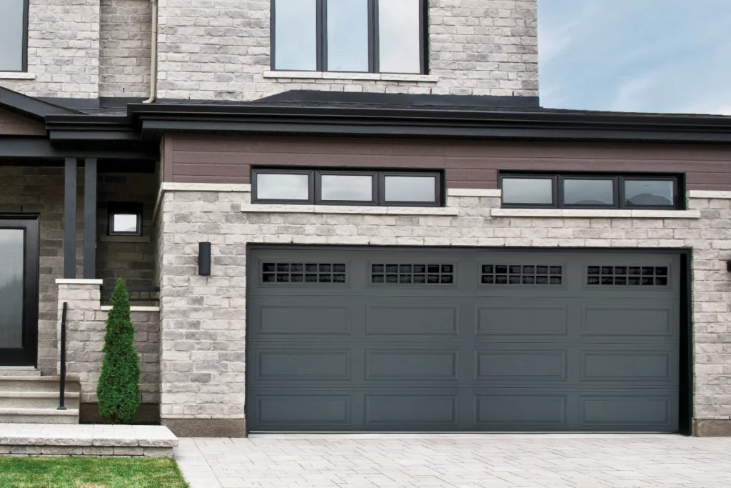 Front of a modern house with a gray garage door, windows above, brick and wood siding, and a small shrub next to steps leading to the entrance.