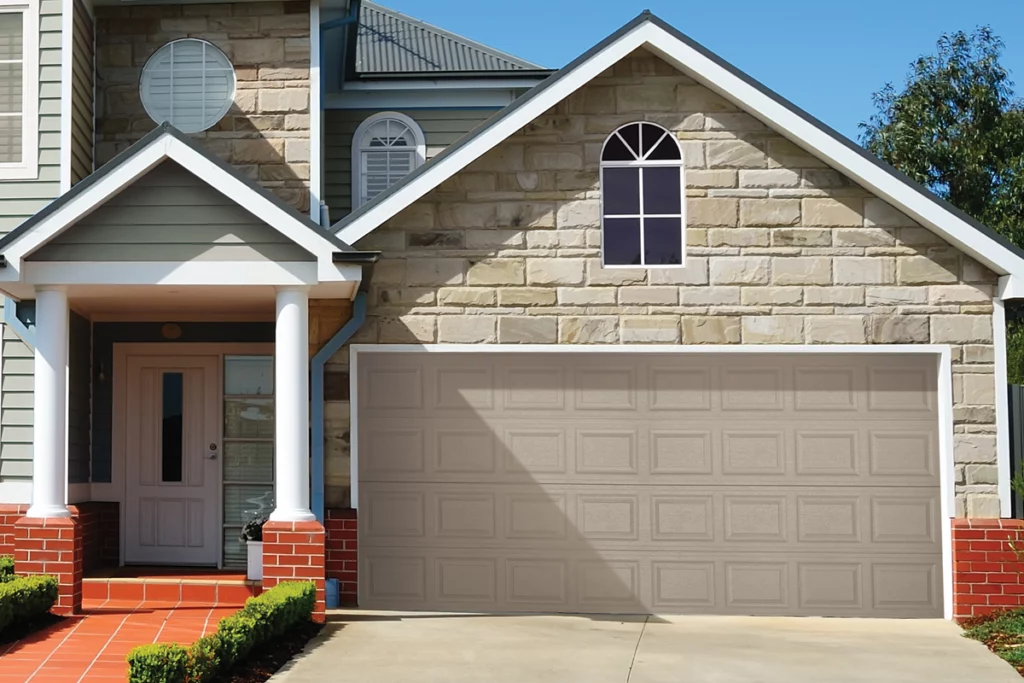 Front view of a house with a beige garage door, stone and vinyl siding, a small porch with white columns, and brick stairs. There are two small bushes along the pathway to the porch and a tree on the right side.