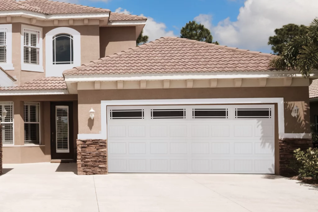 Front of a two-story house with a beige exterior, white windows with shutters, and a white garage door. The roof is tiled with pinkish-brown shingles, and there are some trees and a partly cloudy sky in the background.
