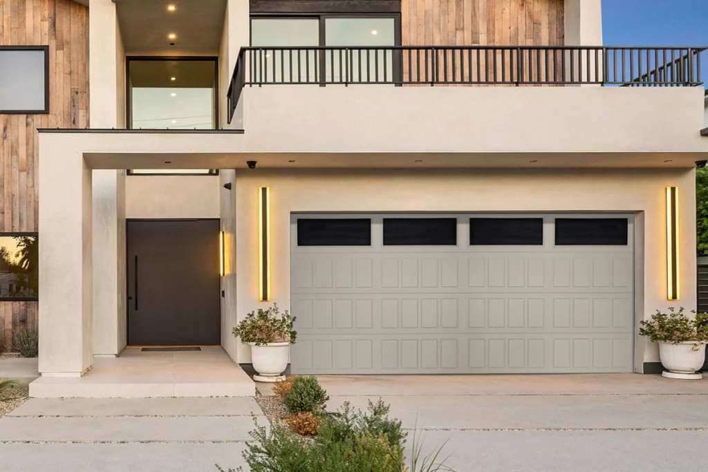 Modern house with a large garage door, front porch, potted plants, and balcony on the upper floor, illuminated by vertical outdoor lights.