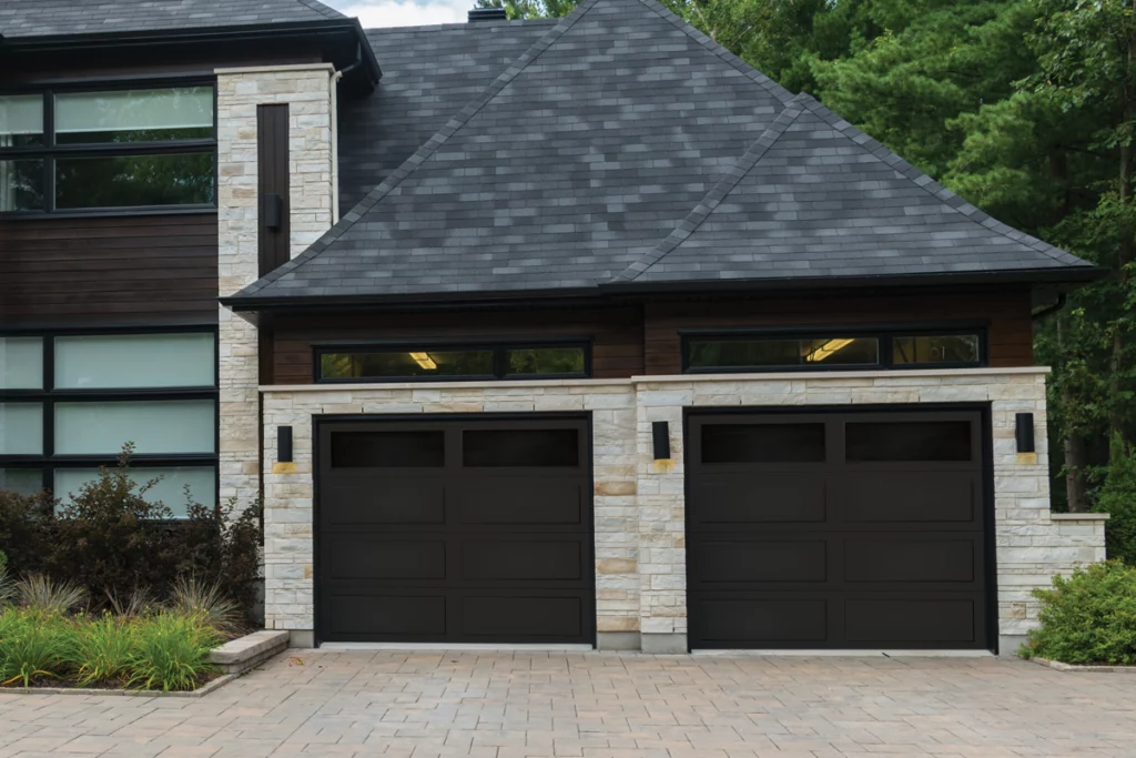 Modern house with stone and wood exterior, black double garage doors, and a paved driveway, surrounded by greenery.
