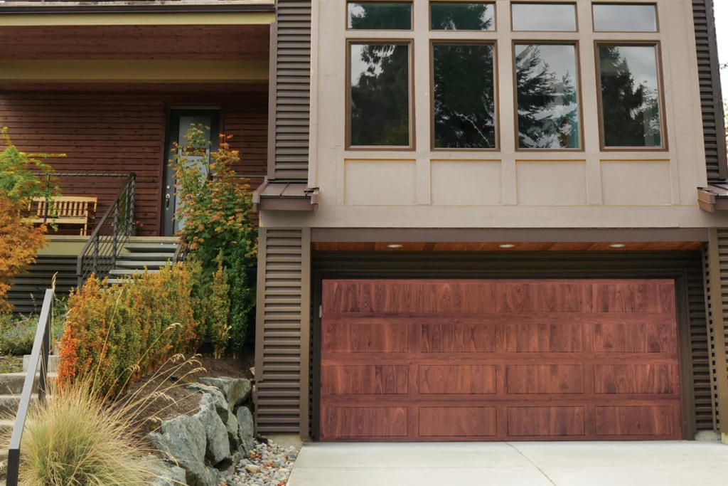 Front view of a modern house with a brown wooden garage door, large windows above, and a small landscaped area with plants and rocks to the side.