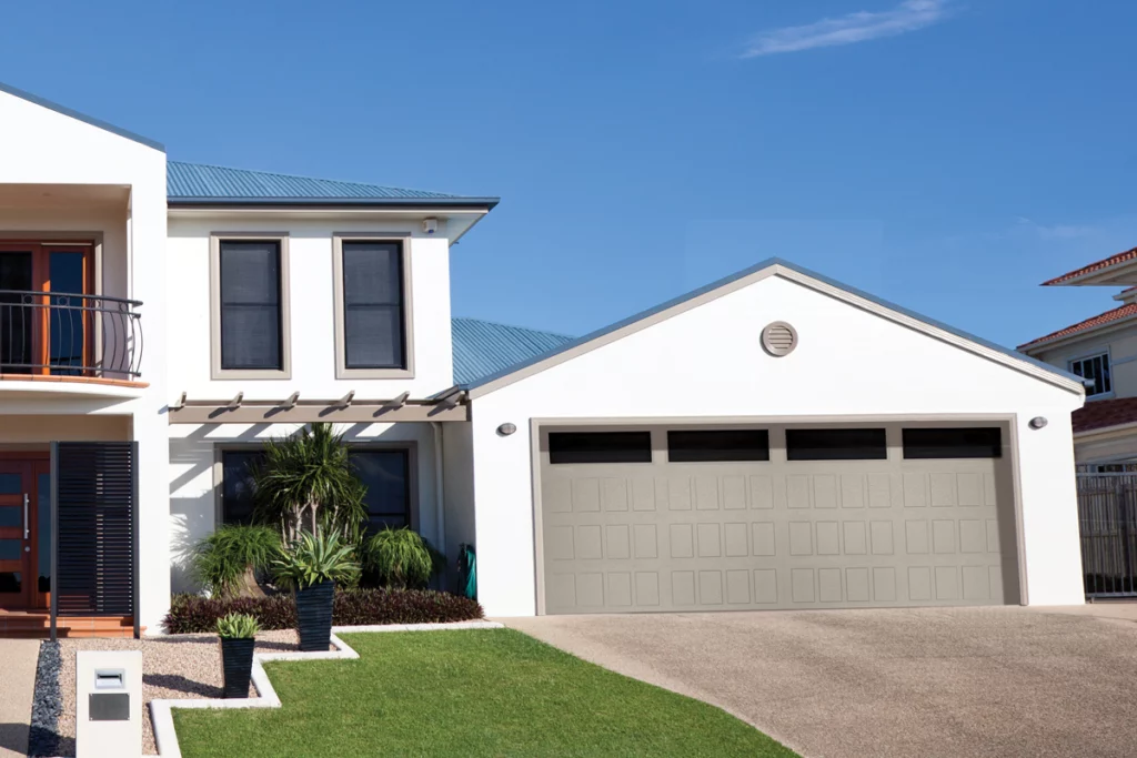 Front view of a modern two-story house with a attached two-car garage, white exterior walls, and a sloped roof, with landscaped front yard including potted plants and a green lawn.