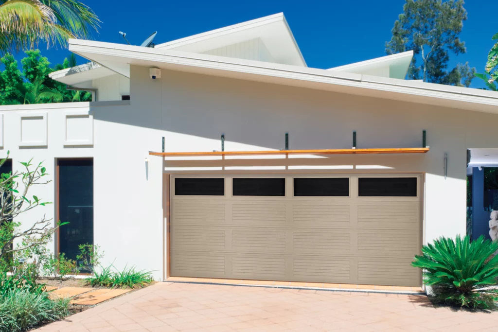 Modern white house with a beige garage door, surrounded by lush green plants, under a blue sky.