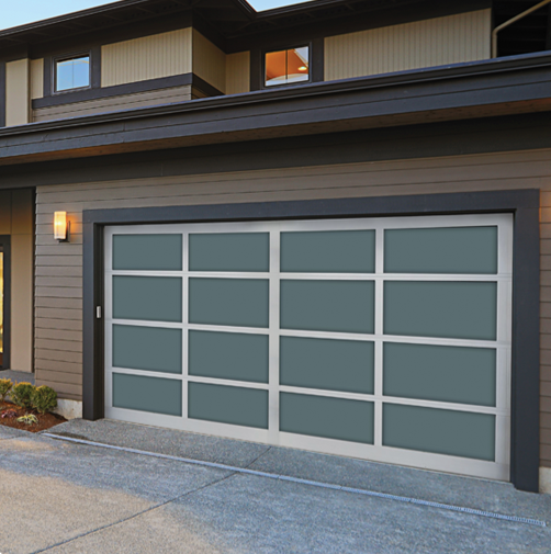 Exterior view of a modern house garage with a closed, gray-and-matte-finish sectional garage door. There is a small light fixture on the gray wall to the left of the garage door, and a concrete driveway in front.