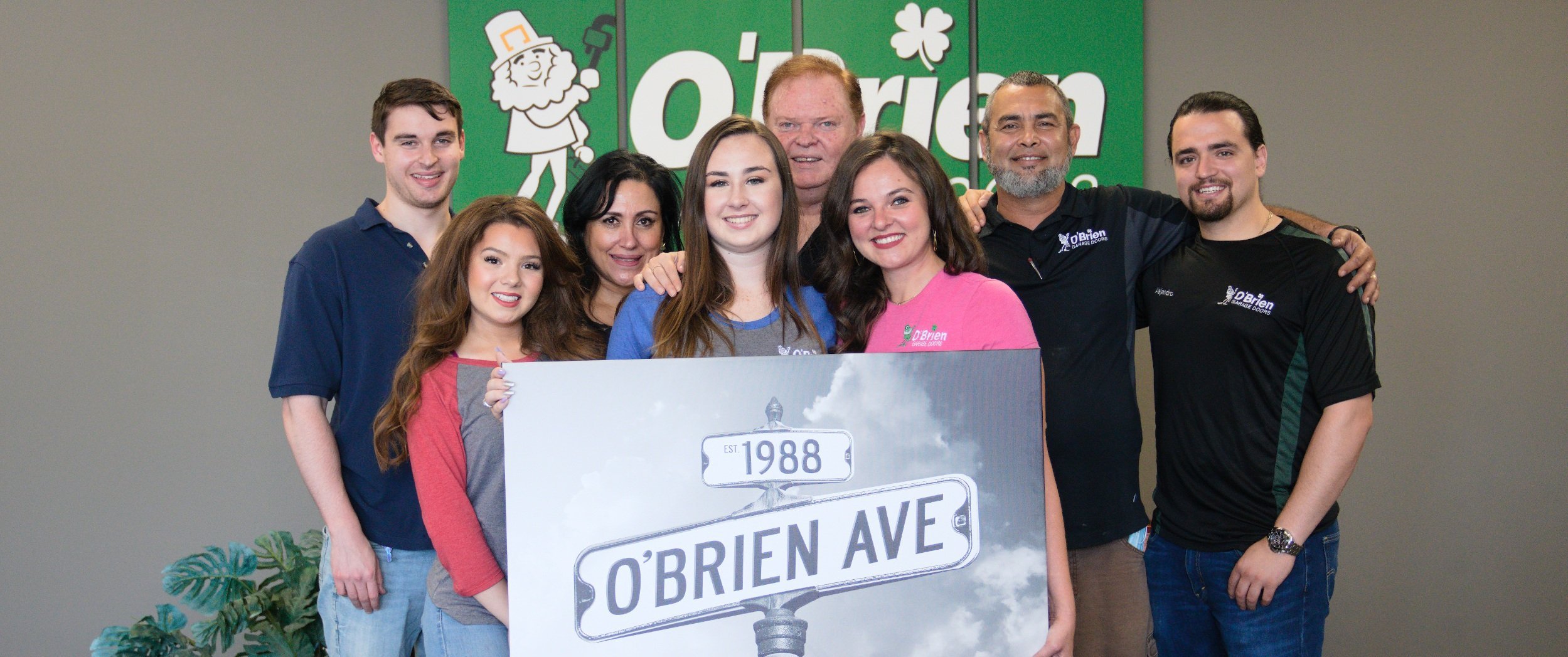 Group of people standing together holding a street sign that reads "O'Brien Ave," at an event for O'Brien Garage Doors, with a green and white logo in the background.