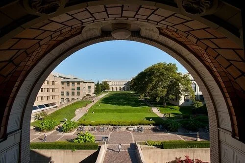 View through an archway of a lush green park with pathways and trees, surrounded by buildings on a sunny day.