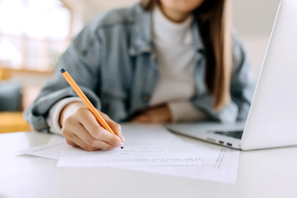 Person writing on paper with a pencil, laptop nearby in a bright room.
