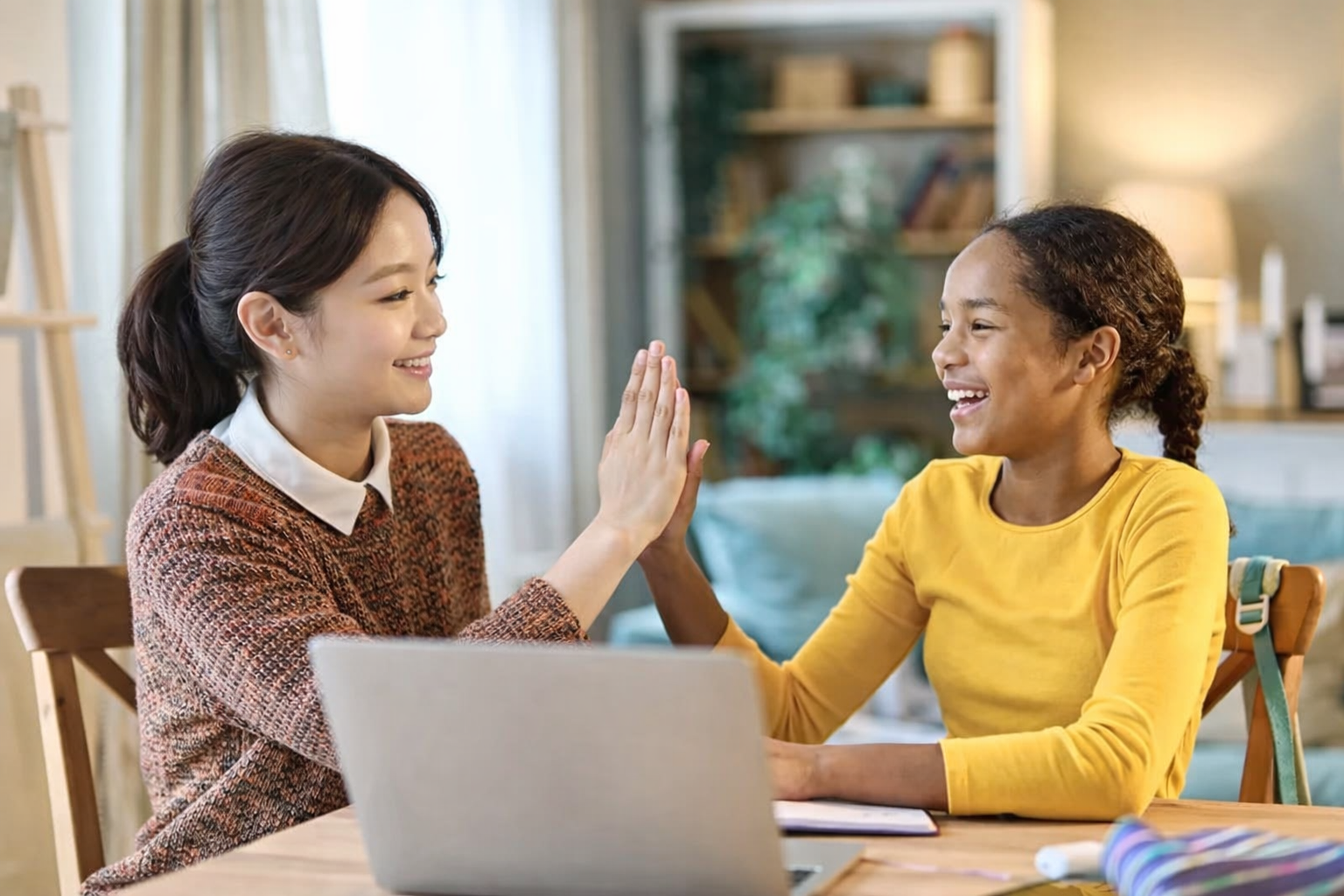 Teacher and student, smiling and high-fiving each other while sitting at a table in a cozy, well-lit room.