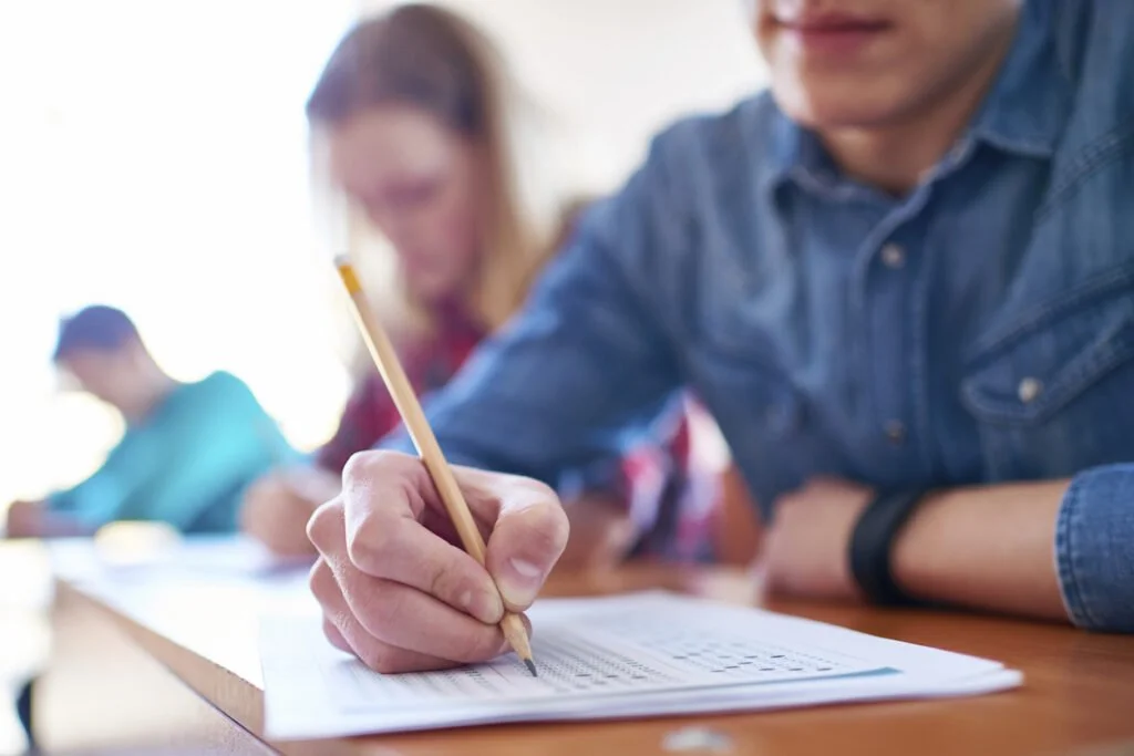 A person holding a pencil and filling out a sheet of paper in a classroom setting, with other students in the background.