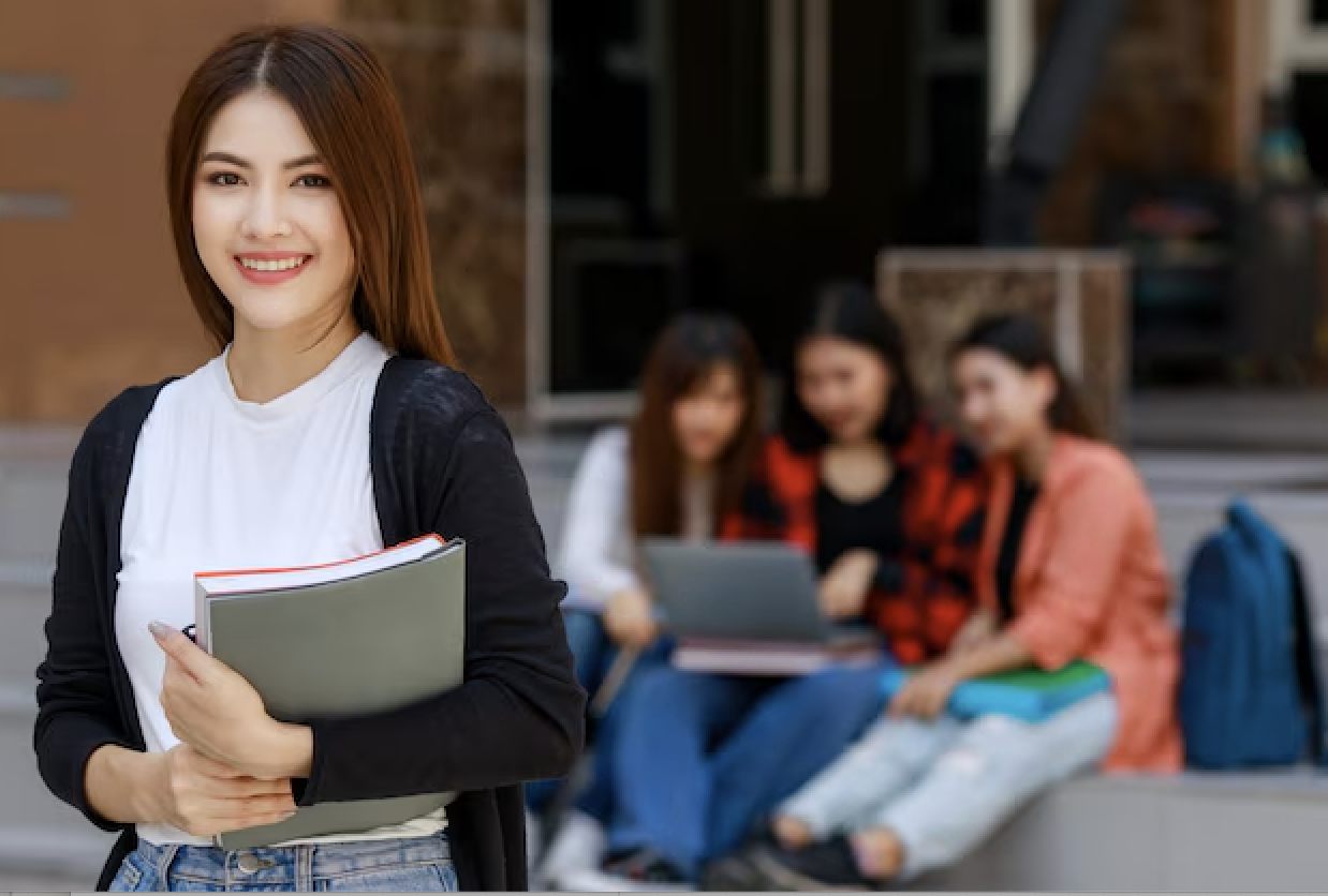A young woman smiling and holding books stands in front of a group of three young women sitting on steps and looking at a laptop.