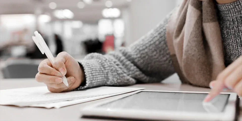 Person writing on paper with a pen while using a tablet in an indoor setting.