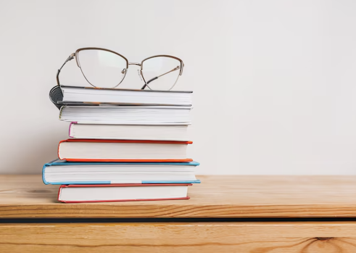 A stack of colorful books topped with a pair of eyeglasses on a wooden surface against a plain white background.