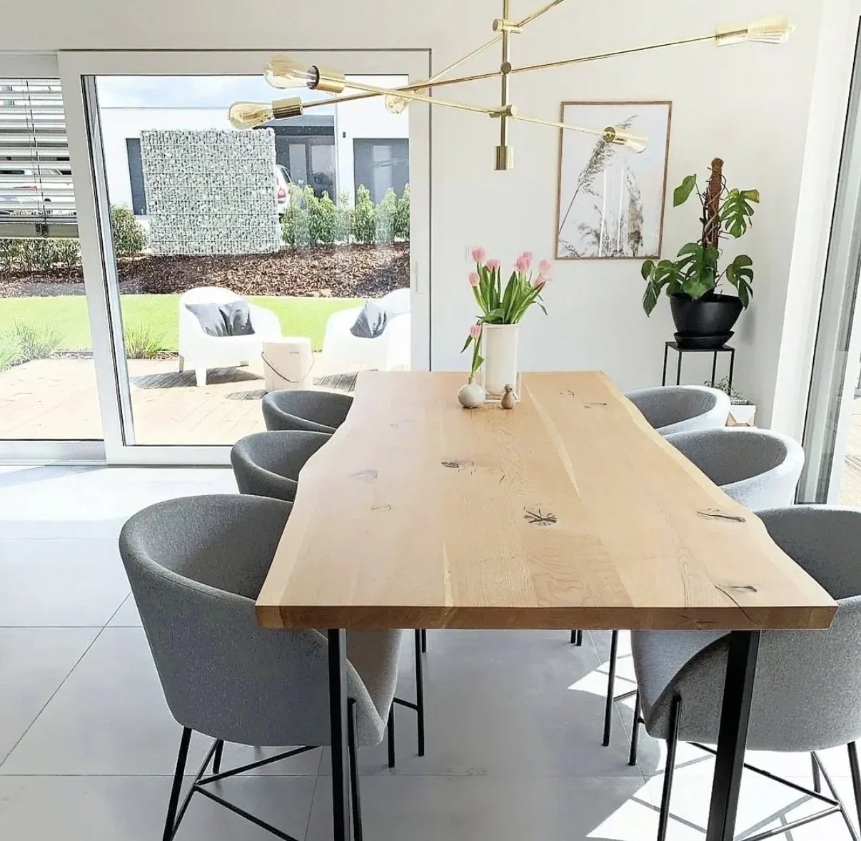 Comedor moderno con una mesa de madera, seis sillas grises, un jarrón con tulipanes rosas y un gran ventanal con vistas a un patio con muebles de exterior y un jardín.