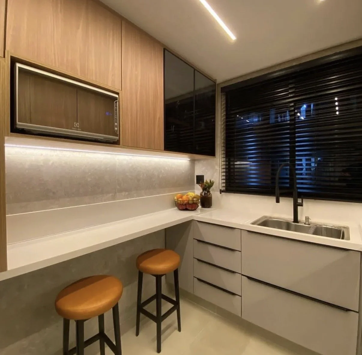 Cocina moderna con armarios de madera y blancos, contraventanas negras, un fregadero, una cesta de fruta y dos taburetes marrones junto a una encimera blanca.