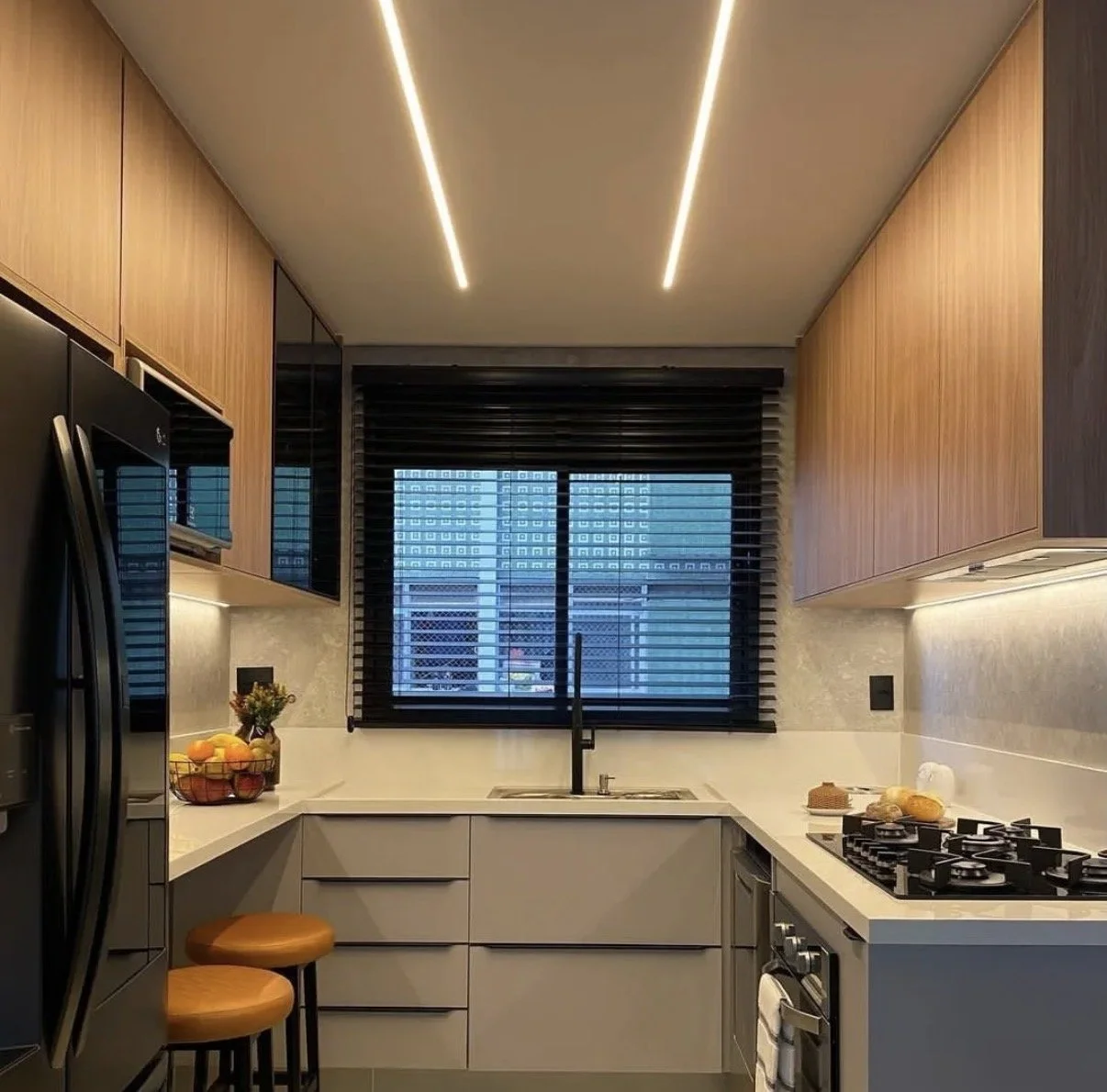 Cocina moderna con frigorífico negro, microondas empotrado, encimeras blancas, armarios superiores de madera, persianas negras y vistas a los edificios de la ciudad.