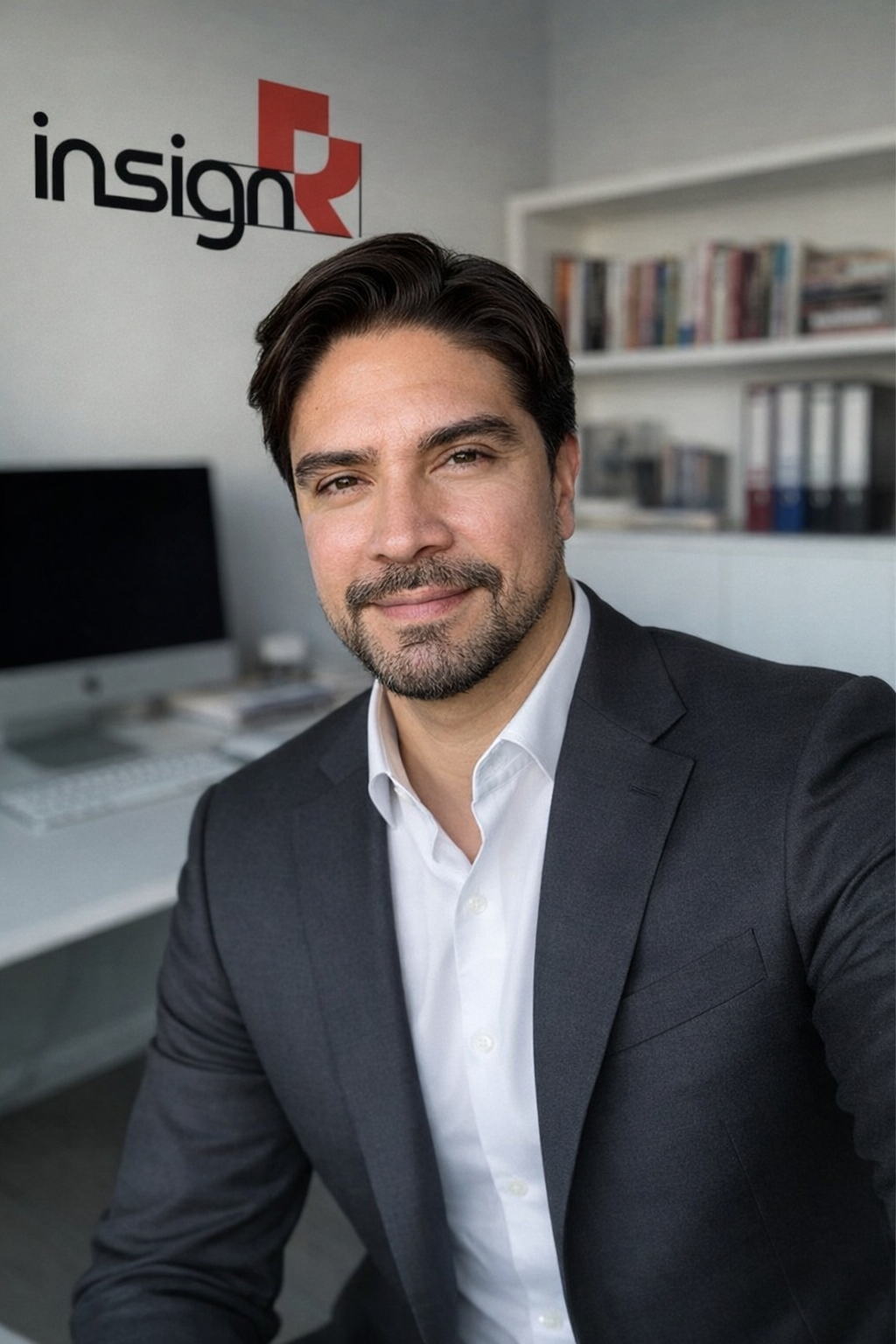 Un hombre vestido con traje haciéndose un selfi en una oficina con un ordenador y una estantería. En la pared hay un logotipo en el que se lee «insign», con un diseño geométrico rojo.