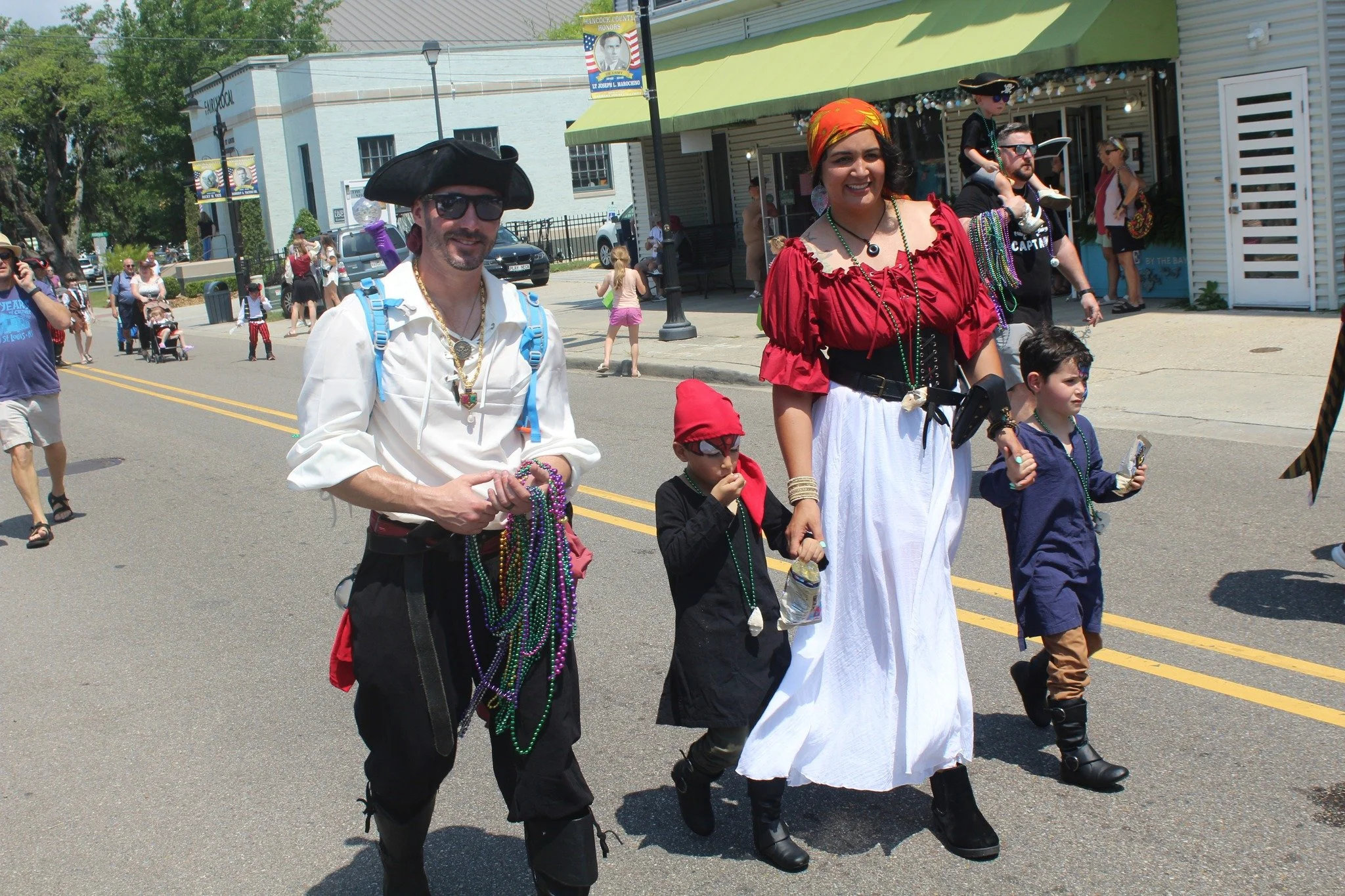 People participating in a parade dressed as pirates, including a man in a black pirate hat and sunglasses, a woman in a red and white pirate costume, and children with pirate accessories.