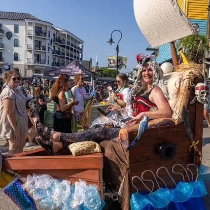 Person dressed as a mermaid sitting on a float with crowds of people around at an outdoor event or festival.