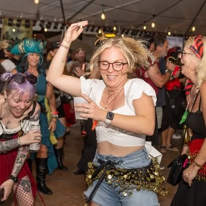 People dancing and having fun at a party, with one woman in the foreground wearing glasses, a white crop top, and a dance belt, surrounded by others in colorful costumes.