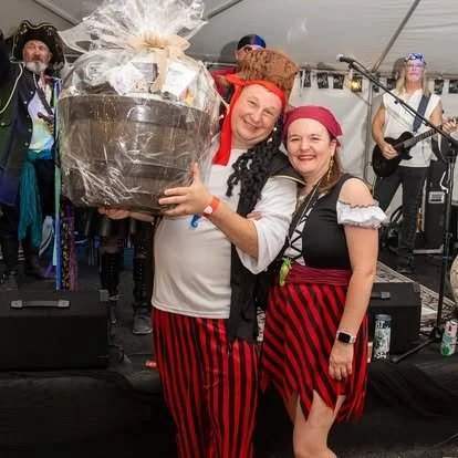 Two women in pirate costumes posing with a large gift basket wrapped in cellophane at a festive event with a band performing in the background.