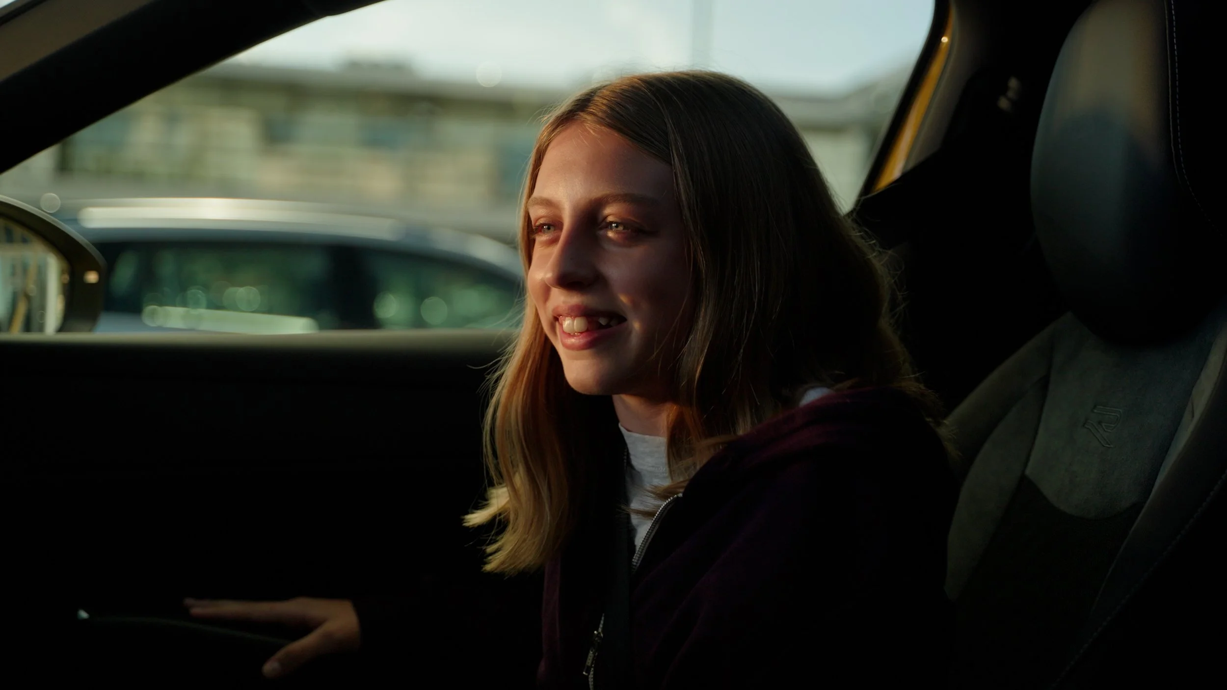 Young girl sitting in a car, smiling and looking outside, with sunlight illuminating her face.