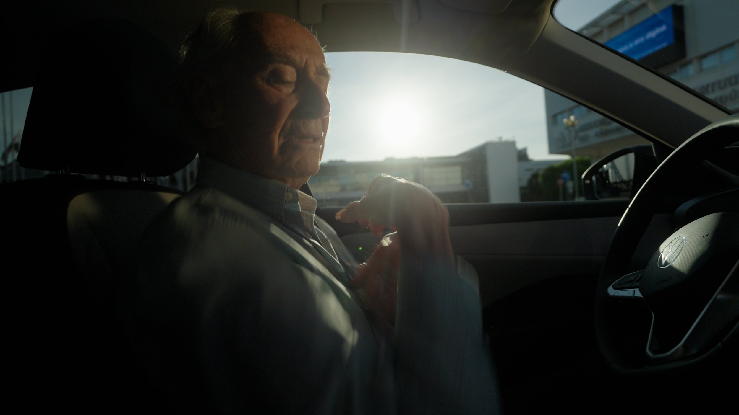 A man sitting in the driver's seat of a car with sunlight shining through the window, adjusting his sleeve or cuff.