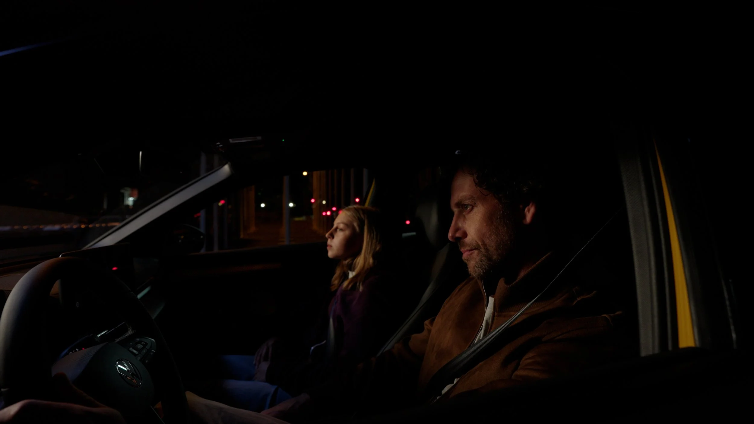 A man and a girl sitting in the front seats of a car at night, with city lights visible through the windows.