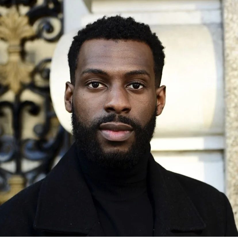 A young Black man with short curly hair and a full beard, wearing a black turtleneck and coat, standing outdoors in front of a decorative wrought iron fence and building.