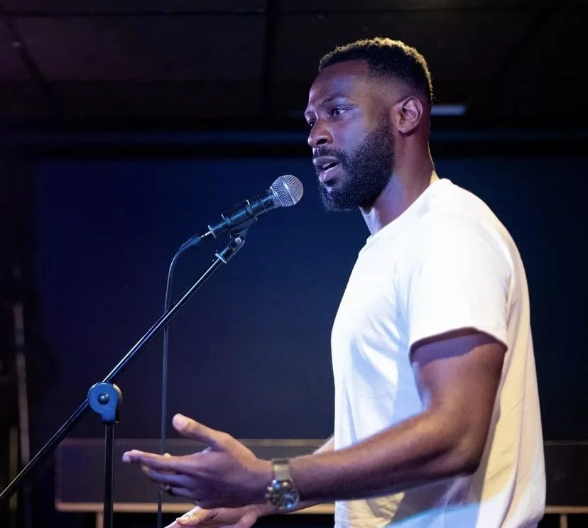 Man speaking into a microphone during a performance or speech, wearing a white t-shirt, with a dark background.