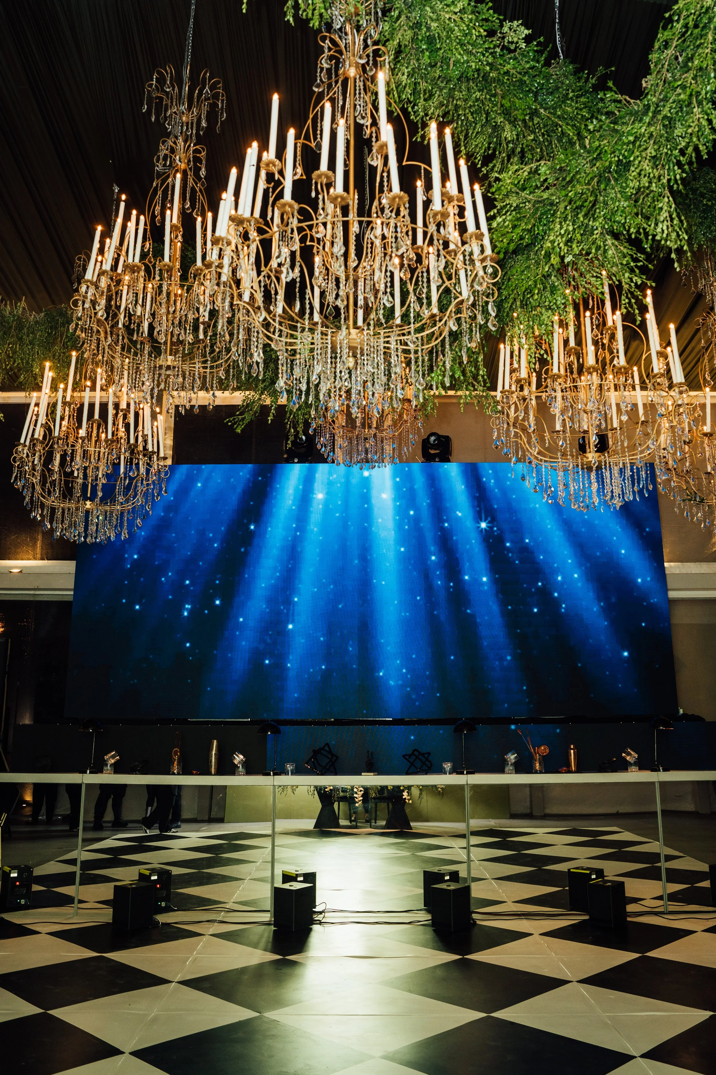 Elegant event space with multiple gold chandeliers decorated with crystals hanging above, a large digital screen displaying a blue starry backdrop, and a black and white checkered floor.
