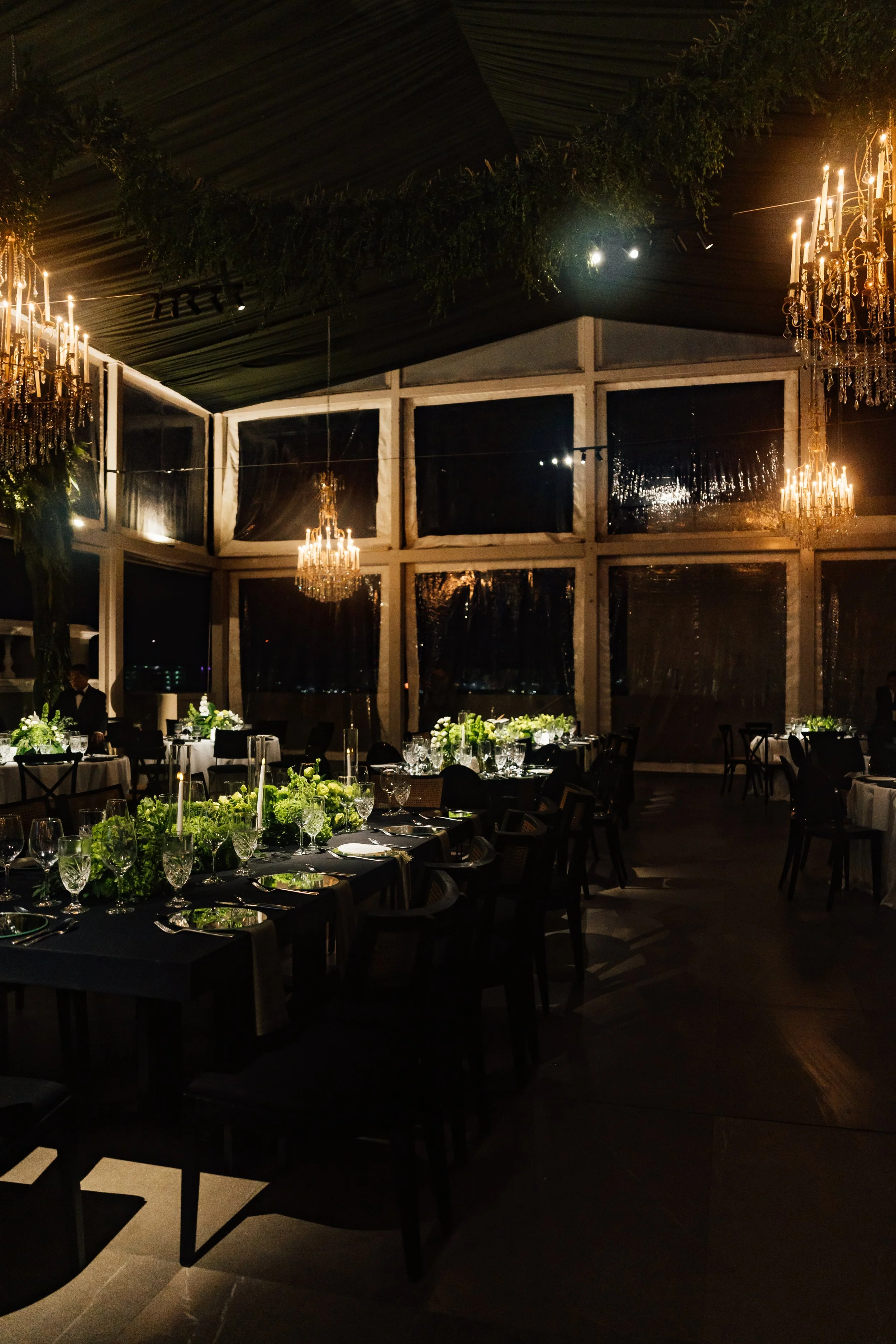 Elegant dinner setup with black tablecloths, green floral centerpieces, and chandeliers in a dimly lit glass-enclosed event space.