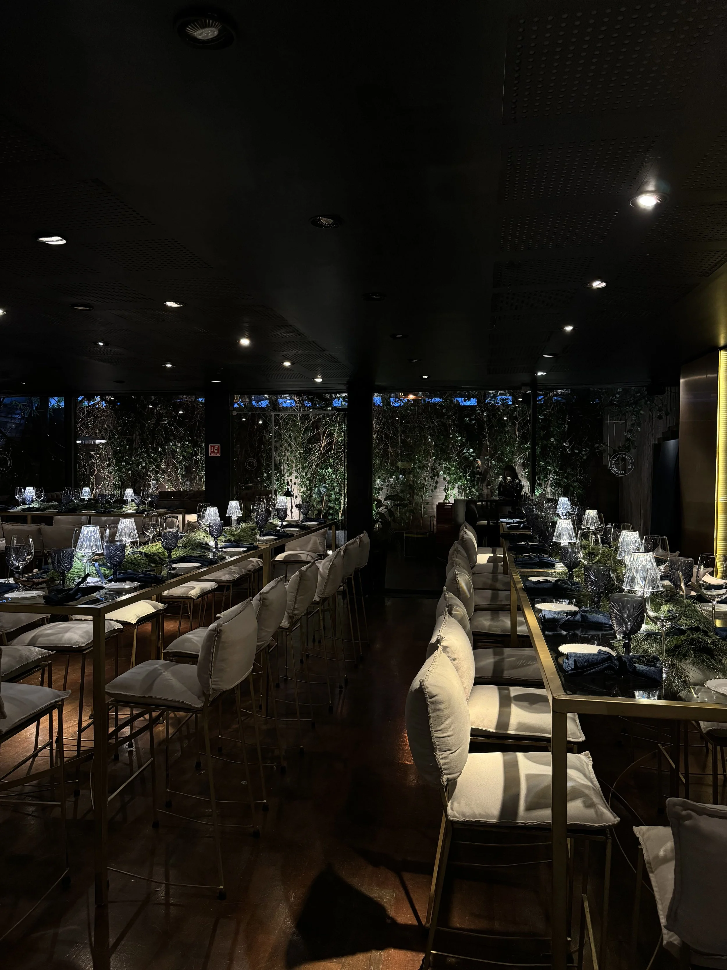 A dimly lit restaurant interior with long dining tables set with wine glasses, dark napkins, and greenery, surrounded by cushioned chairs, and a lush green garden view in the background.
