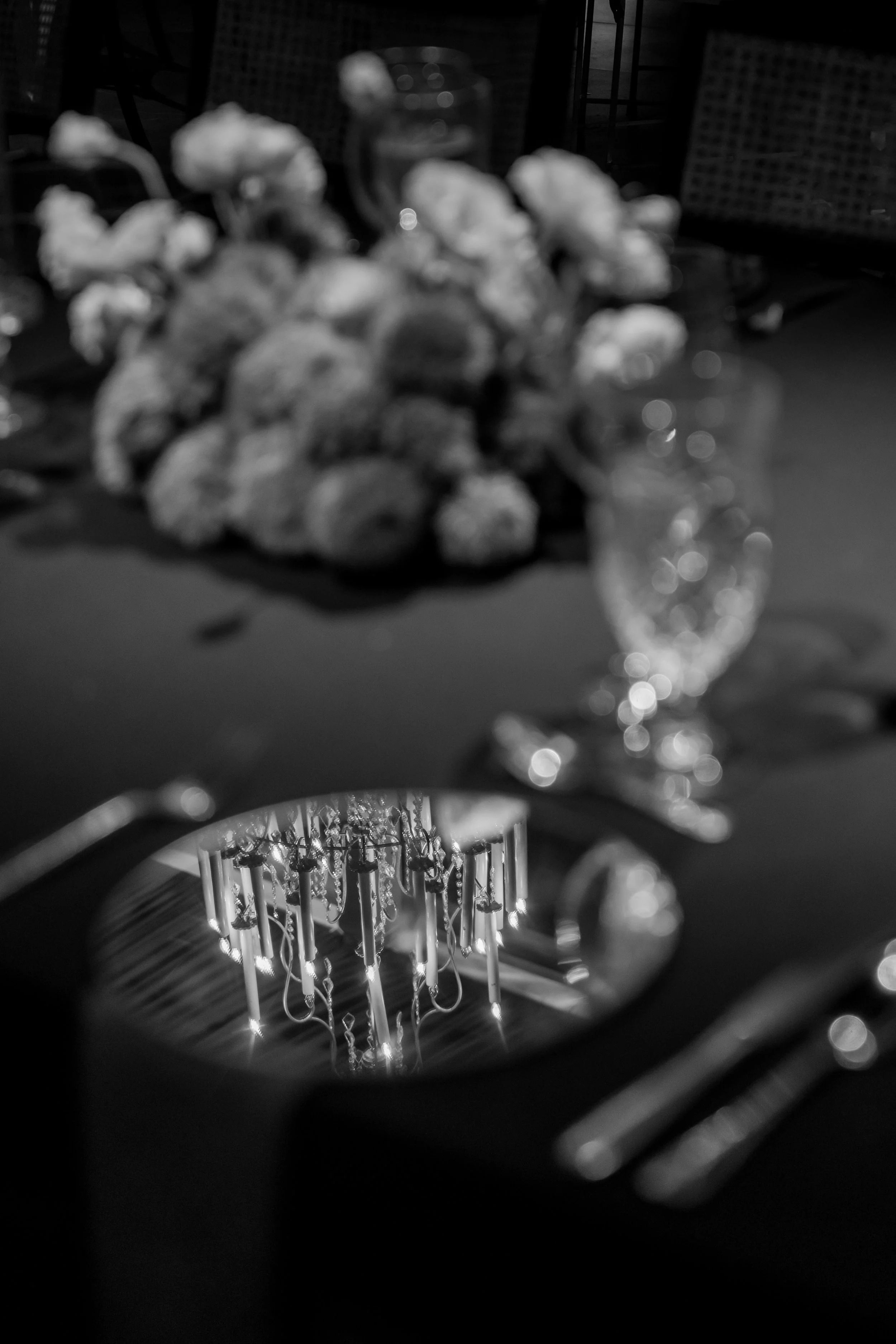 Reflection of a chandelier in a hand mirror on a dining table, with blurred flowers and glasses in the background.