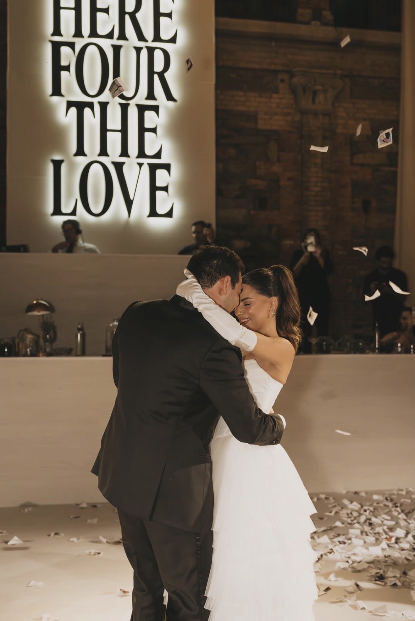 A couple dancing at their wedding reception, with a large illuminated sign in the background reading 'Here four the Love' and confetti on the floor.