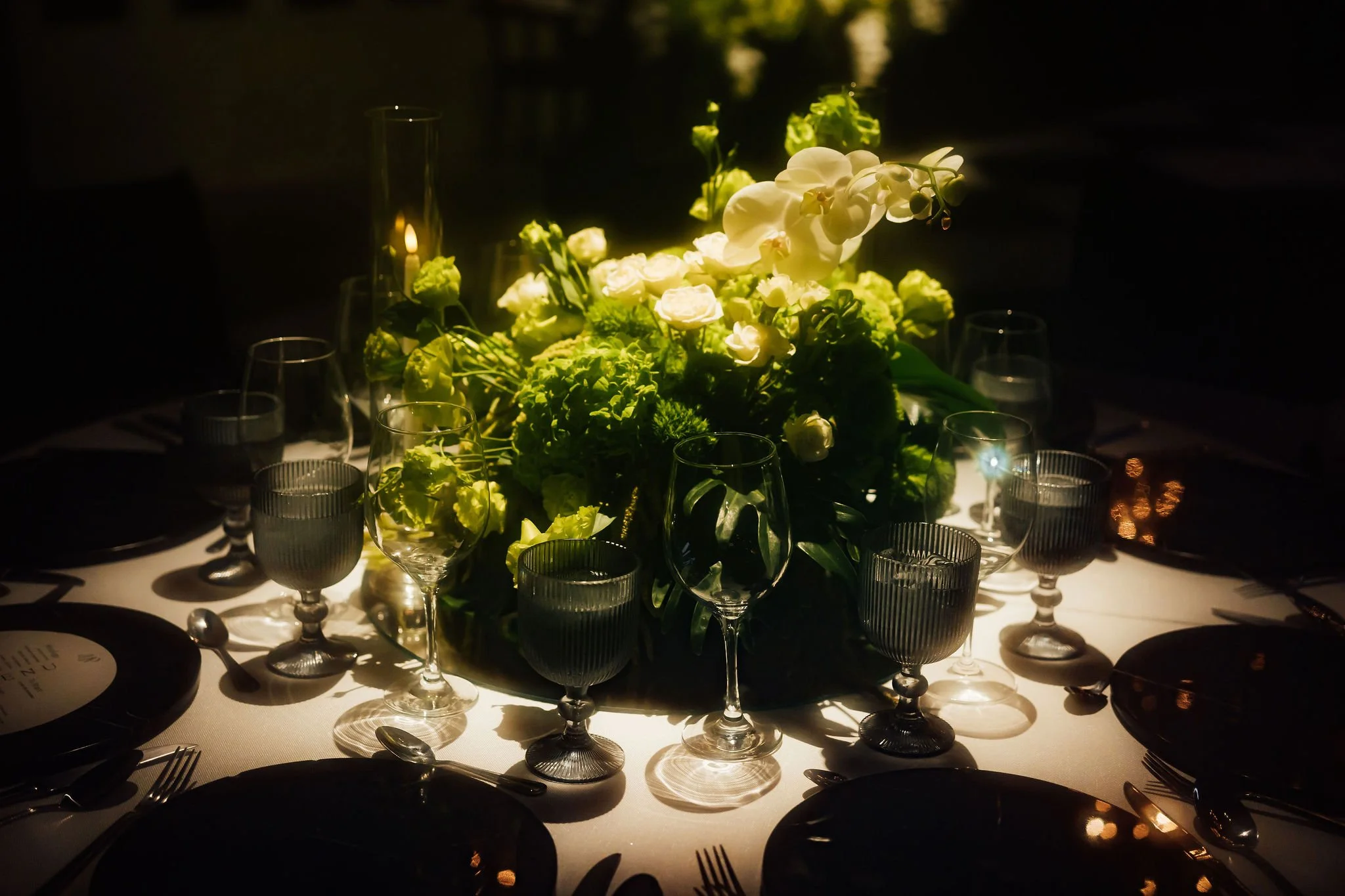 A round dining table set with black plates, silverware, and glasses, featuring a large floral centerpiece with white flowers and green foliage, illuminated softly in a dark setting with candles and reflections.