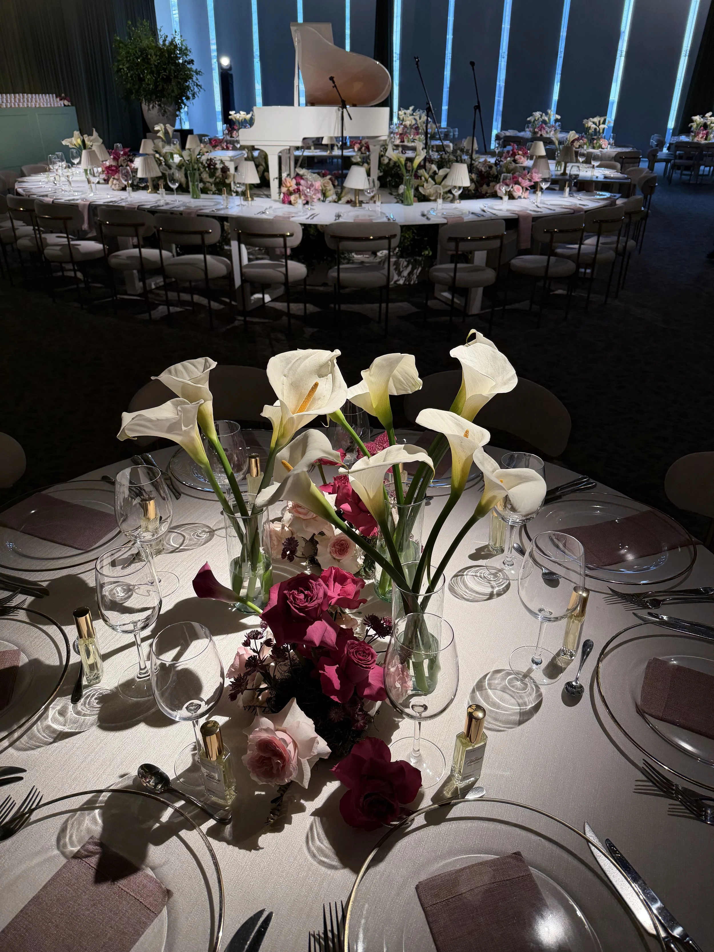 Elegant banquet table set up with floral centerpieces and white tableware, with a grand white piano in the background, in a dimly lit event space.
