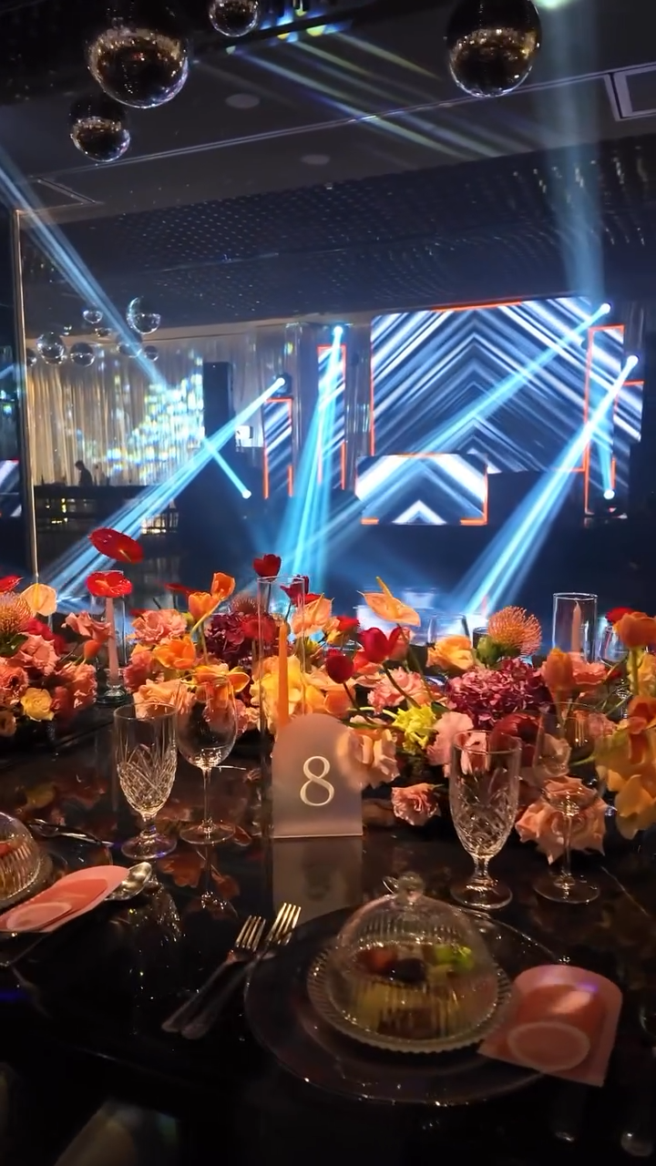 A decorated banquet table with floral arrangements, glassware, and place settings in a dimly lit event space with a stage featuring bright laser lights and geometric patterns.