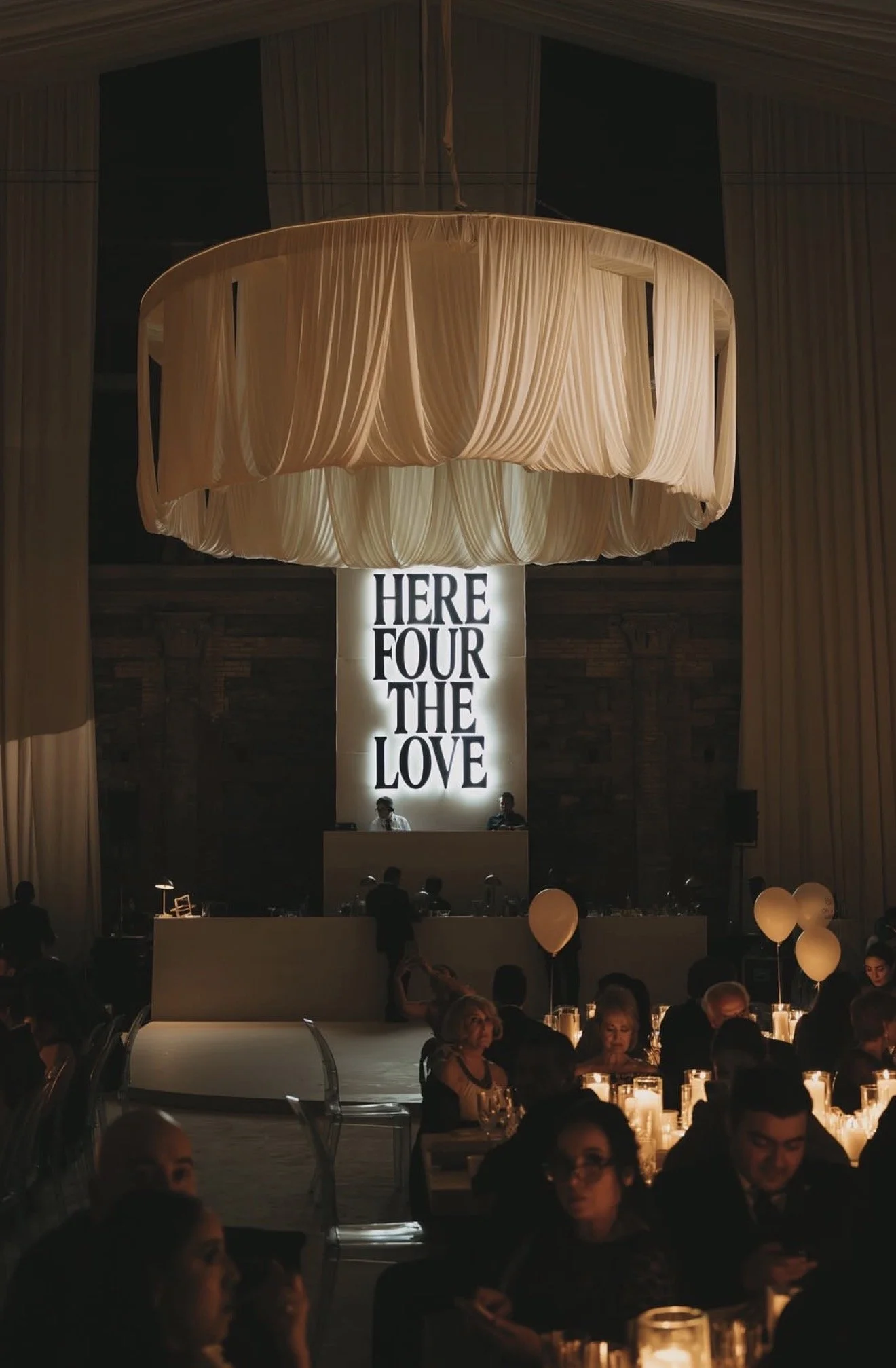 Indoor event with dim lighting, wooden walls, large beige fabric chandelier, illuminated sign that reads 'HERE FOUR THE LOVE', and people seated at tables with candles and balloons, likely a celebration or party.