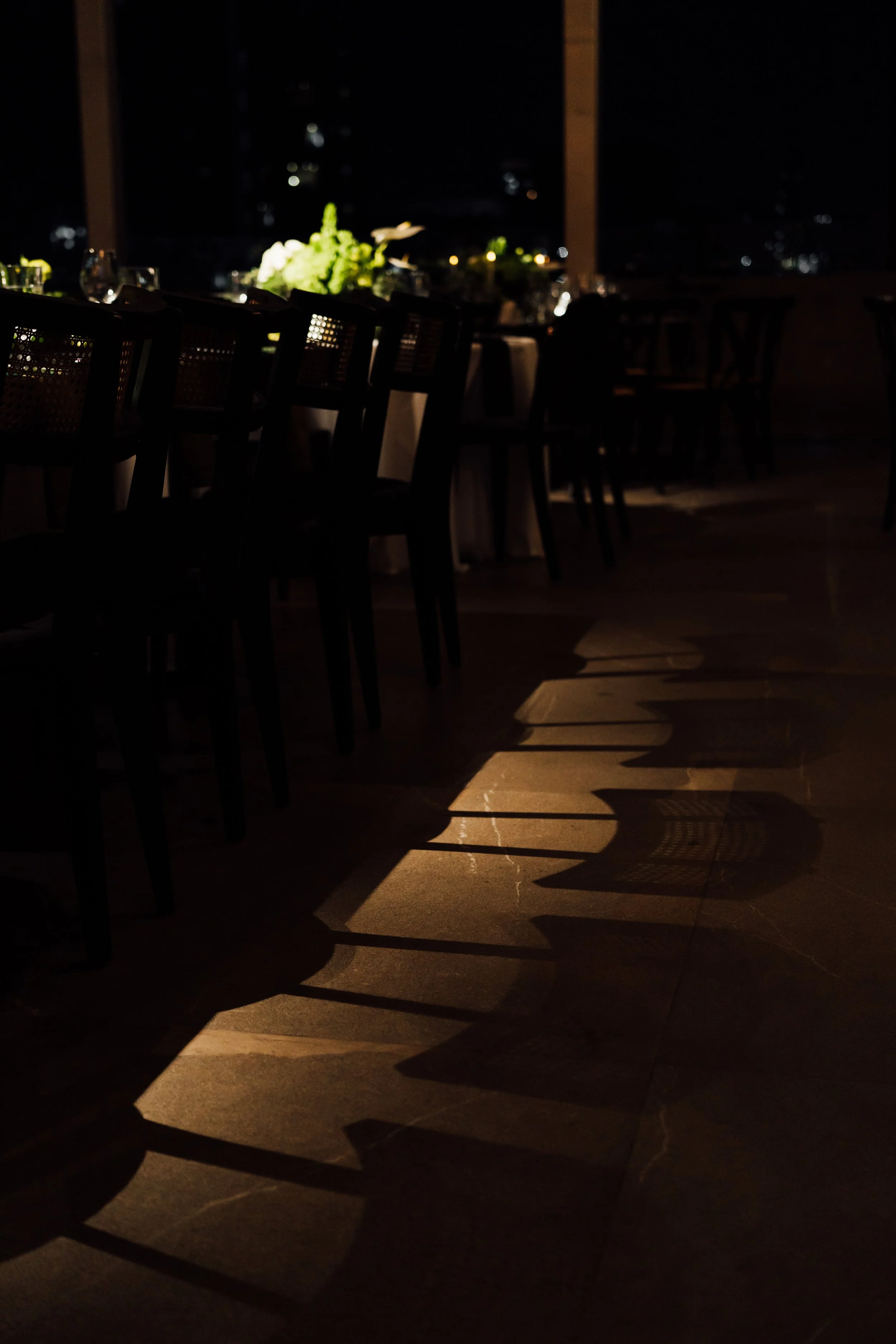 Dimly lit dining area with chairs and a bouquet of flowers on a table, shadowed against large windows showing city lights at night.
