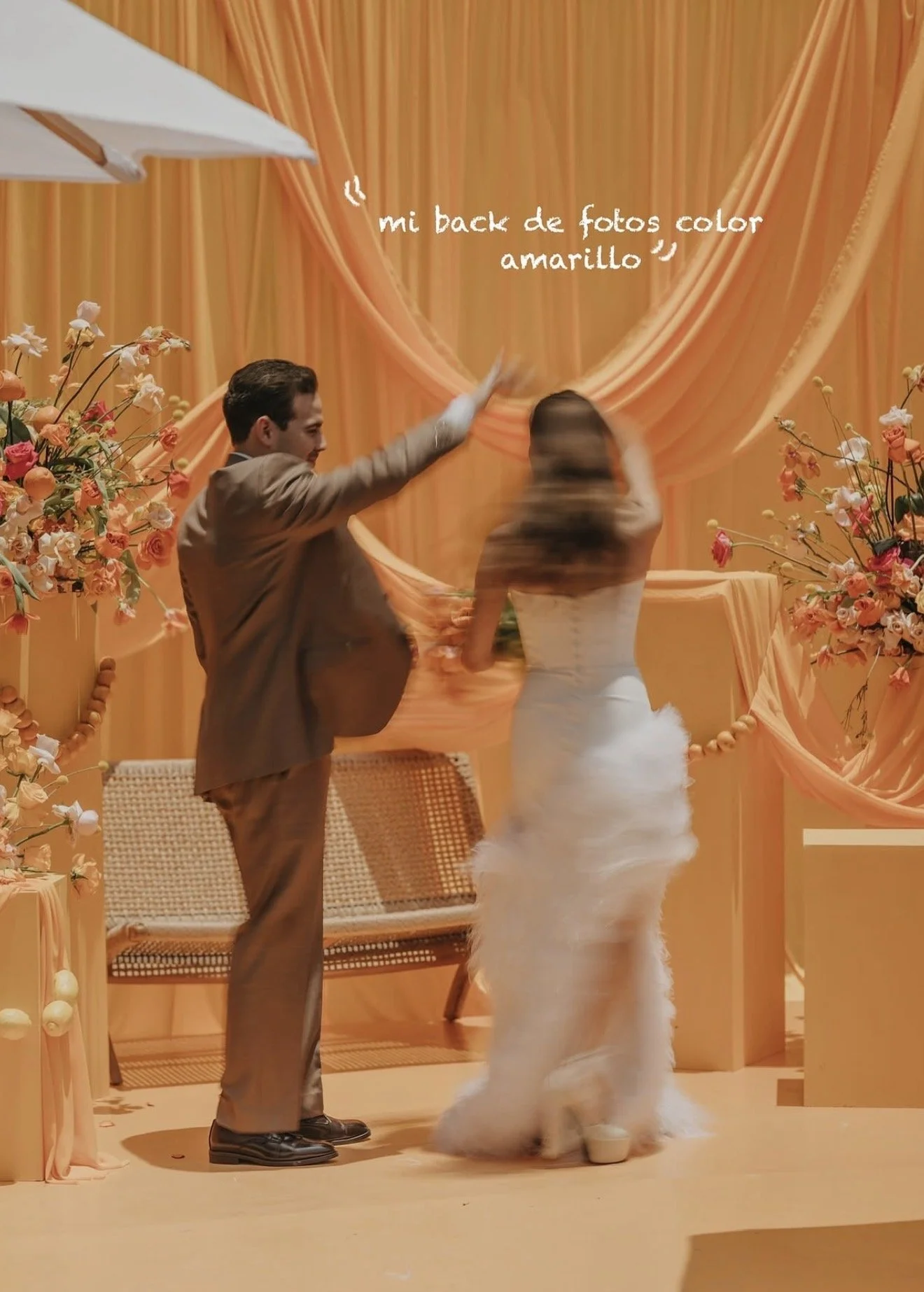 A man and a woman at a wedding reception or celebration with a yellow or orange background, floral decorations, and a bench.