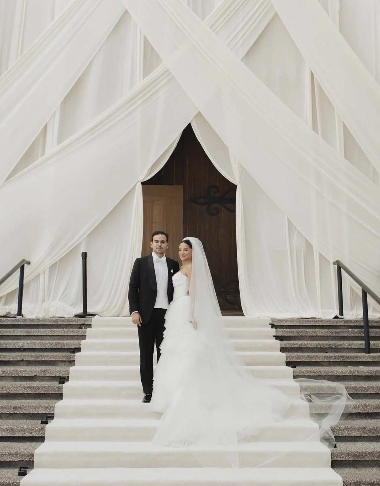 A bride and groom standing on white steps inside a church, with draped white fabric and wooden walls in the background.