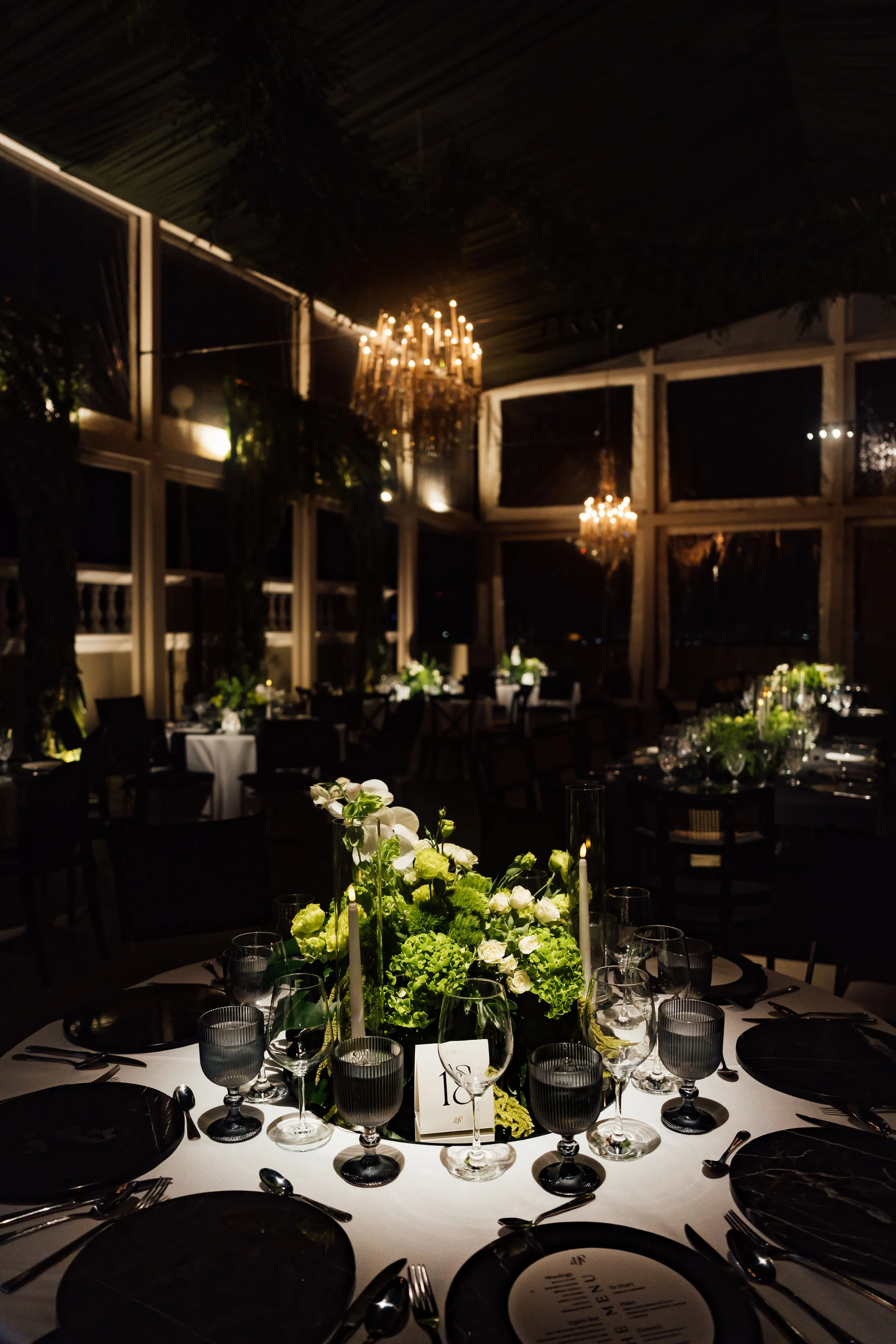 A decorated banquet table with black plates, glassware, and floral centerpiece at an elegant evening event in a dimly lit room with chandeliers and large windows.