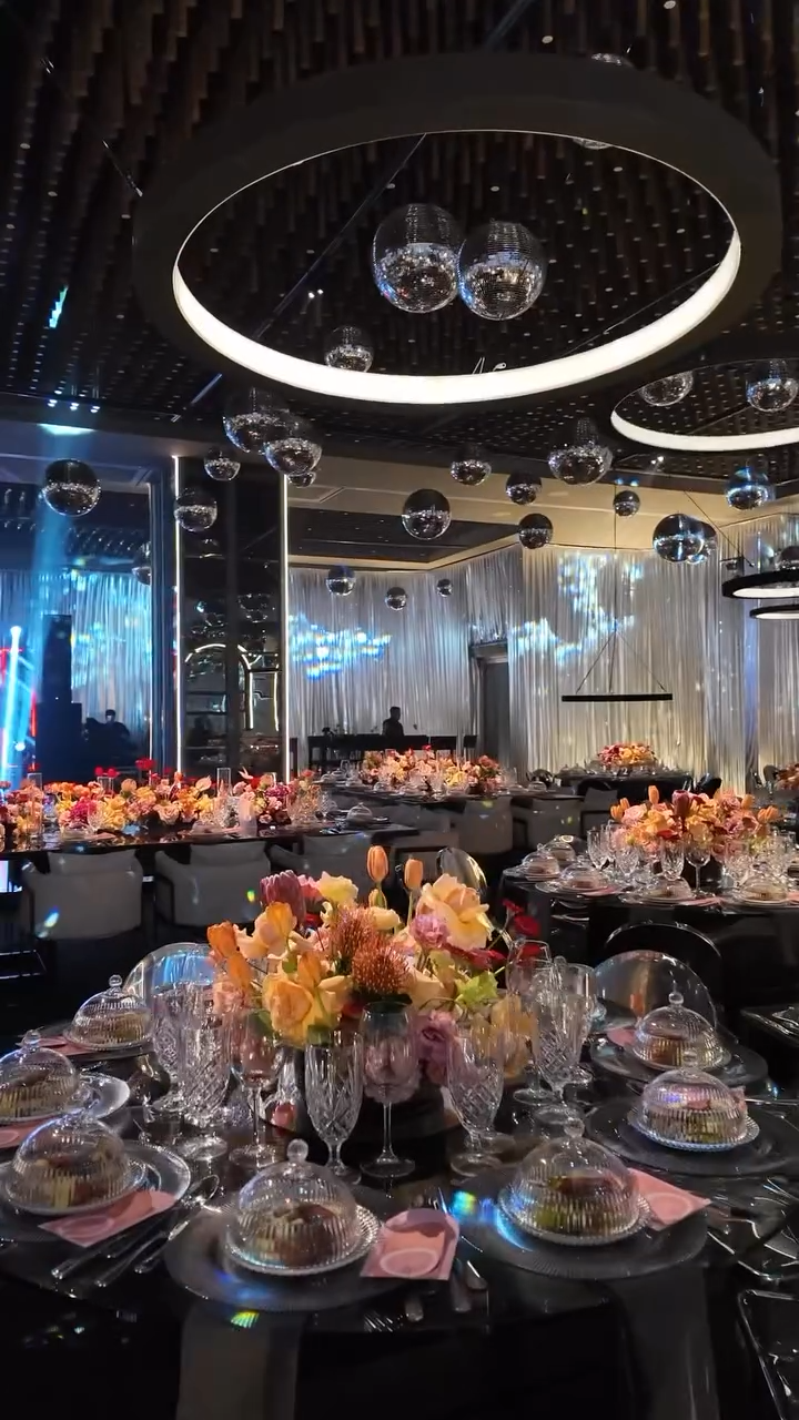 Elegant event venue decorated with floral centerpieces, top glasses with cakes underneath, mirrored disco balls, and modern ceiling lights, set for a banquet or celebration.