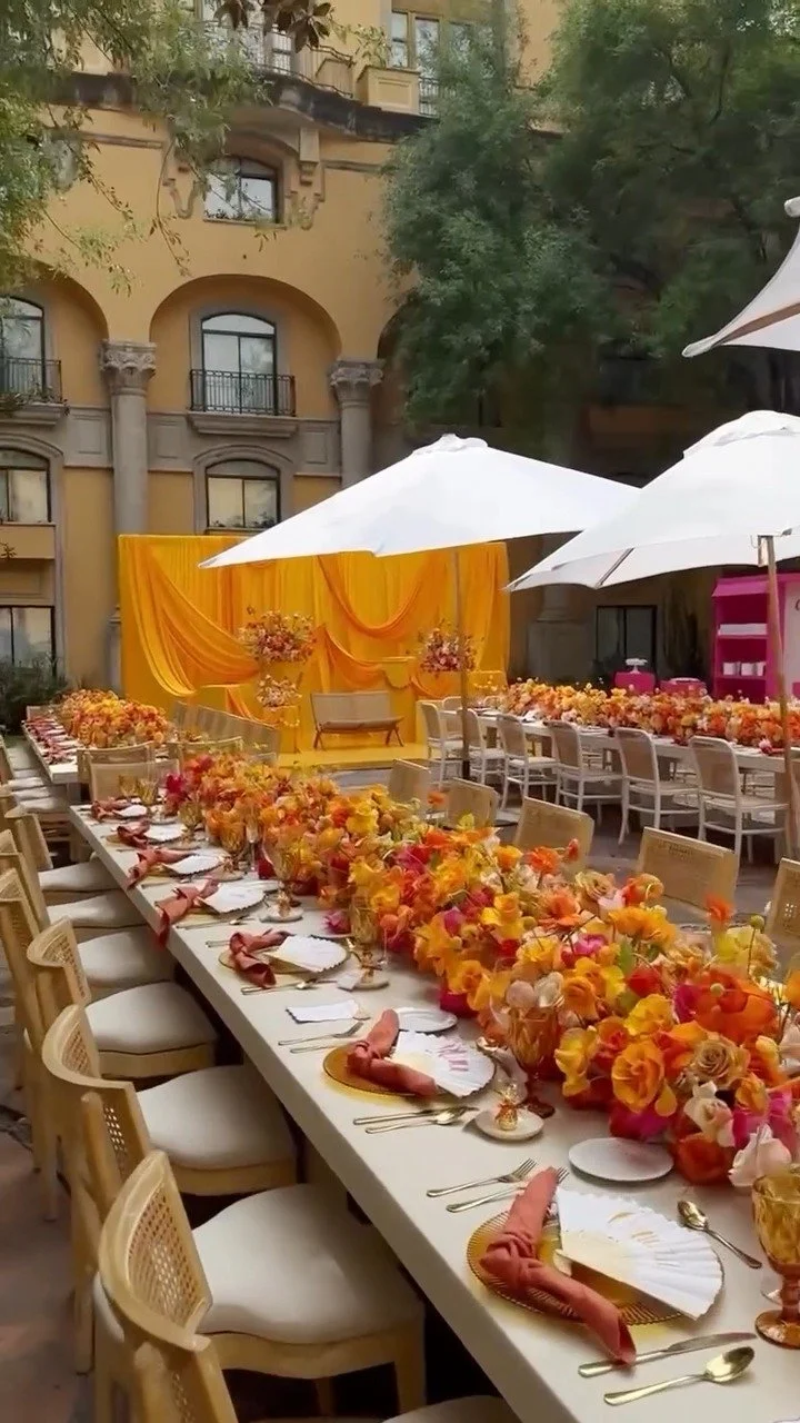 Long rectangular table set for a celebration with colorful floral arrangements, gold chargers, peach napkins, and white plates, under large white umbrellas, with a yellow draped backdrop and an ornate building in the background.