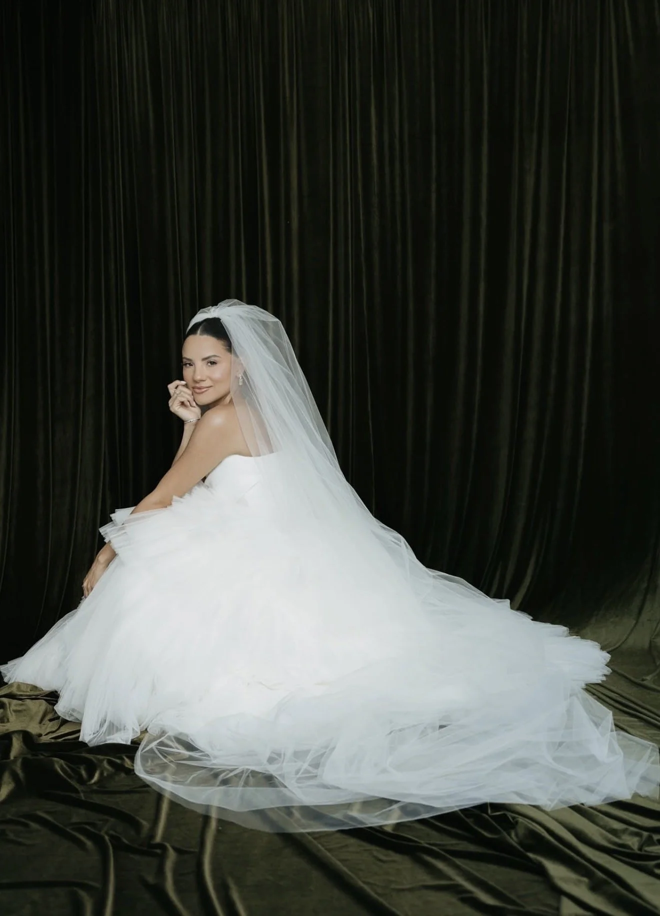 Bride in a wedding dress sitting on a chair with a white veil and a black curtain background.